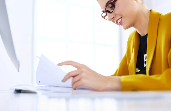 Красивая молодая деловая женщина, занимающаяся бумажной работой, сидя за рабочим столом перед ноутбуком — стоковое фото