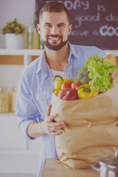 Uomo in possesso di sacchetto di carta pieno di generi alimentari sullo sfondo della cucina. Shopping e cibo sano concetto — Foto Stock