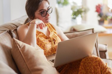 Evde dizüstü bilgisayar üzerinde çalışan kanepede oturan genç kadın