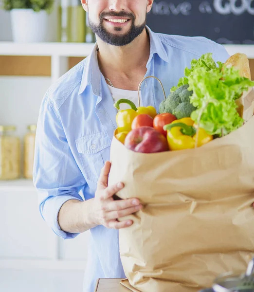 Hombre sosteniendo bolsa de papel llena de comestibles en el fondo de la cocina. Compras y concepto de comida saludable — Foto de Stock