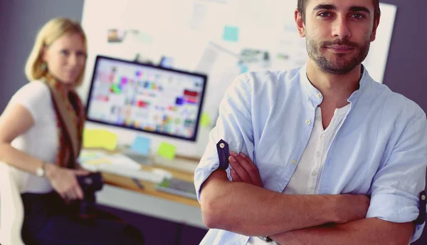 Porträt eines jungen Designers vor Laptop und Computer während der Arbeit. Assistentin bedient ihr Handy im Hintergrund. — Stockfoto