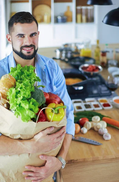 Hombre sosteniendo bolsa de papel llena de comestibles en el fondo de la cocina. Compras y concepto de comida saludable — Foto de Stock