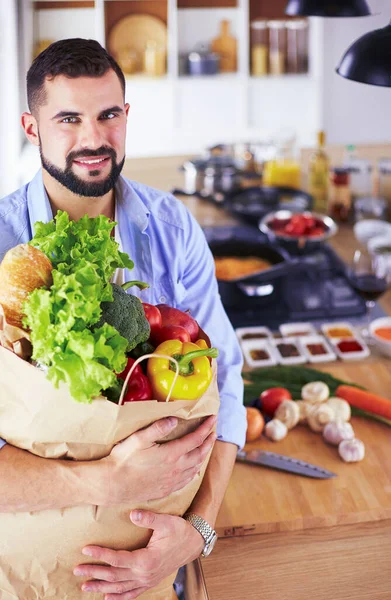 Hombre sosteniendo bolsa de papel llena de comestibles en el fondo de la cocina. Compras y concepto de comida saludable — Foto de Stock