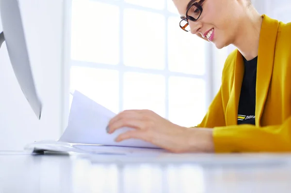 Hermosa joven mujer de negocios haciendo un poco de papeleo mientras está sentado en el escritorio de la oficina delante de la computadora portátil —  Fotos de Stock