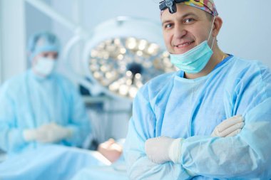 Ameliyat odasında arka planda erkek cerrah var.