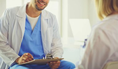 Yakışıklı doktor genç bayan hastasıyla konuşuyor ve ofisinde otururken notlar alıyor.