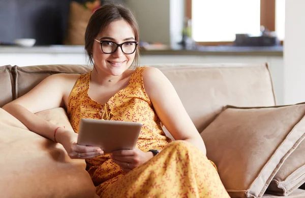 Güzel genç esmer kadın evde kanepe veya kanepe üzerinde onu tablet bilgisayar kullanarak oturan ve gülümseyen — Stok fotoğraf