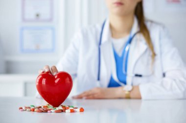 Beyaz önlüklü doktor. Steteskoplu. Elinde haplar ve beyaz bir kalbe takılmış plastik kalp var.