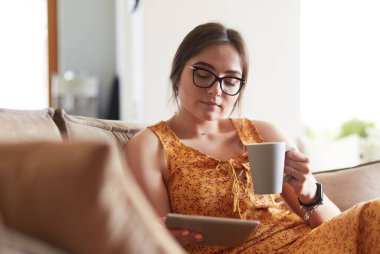Genç kadın evinde kanepede kahve içerken tablet kullanıyor.