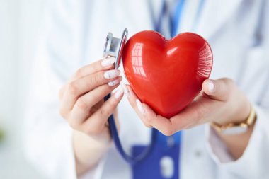 Steteskoplu bir doktor, beyaz arka planda kırmızı kalbi inceliyor.