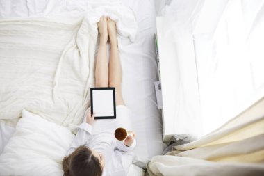 Sabahları yatakta otururken tablet kullanan ve kahve içen kadın portresi