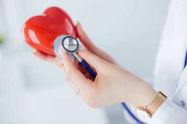 Steteskoplu bir doktor, beyaz arka planda kırmızı kalbi inceliyor.