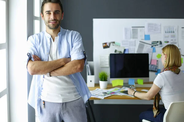 Porträt eines jungen Designers vor Laptop und Computer während der Arbeit. Assistentin bedient ihr Handy im Hintergrund. — Stockfoto