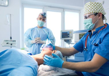 Ameliyat odasında arka planda erkek cerrah var.