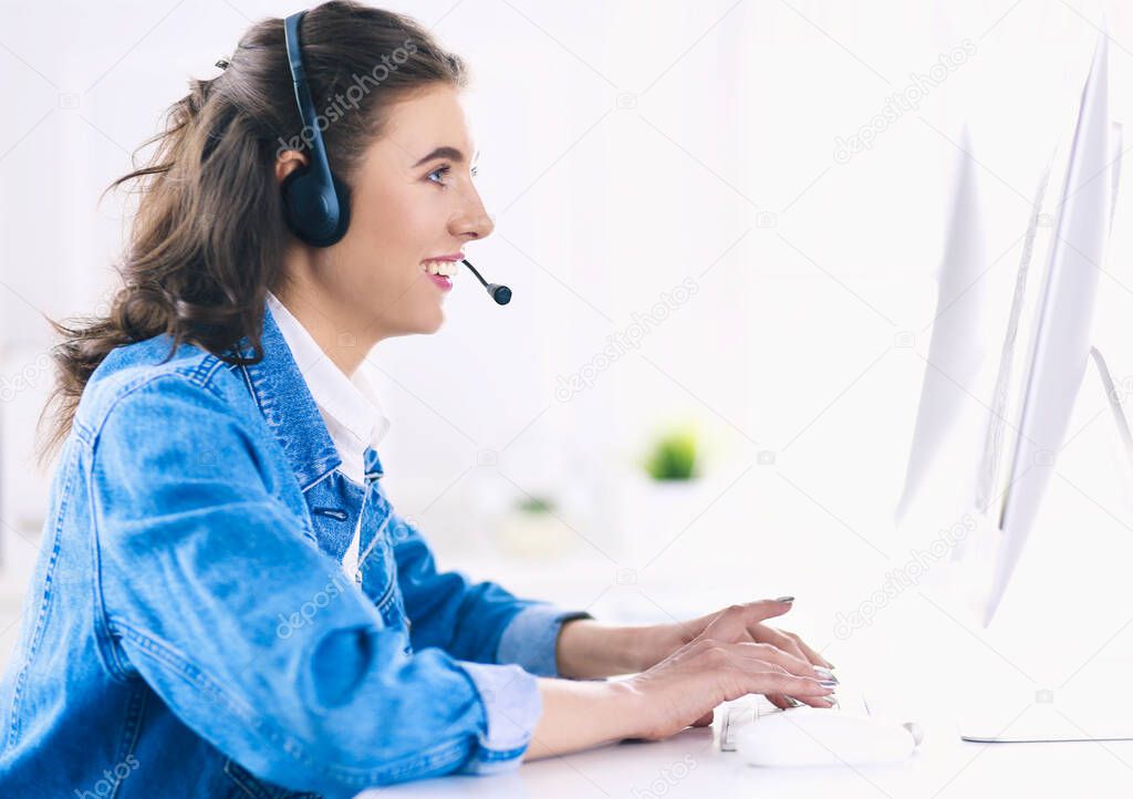 Happy charming young woman sitting and working with laptop using headset in office