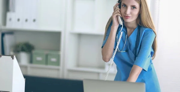 Mladá žena lékař v bílém kabátu na počítači pomocí telefonu — Stock fotografie