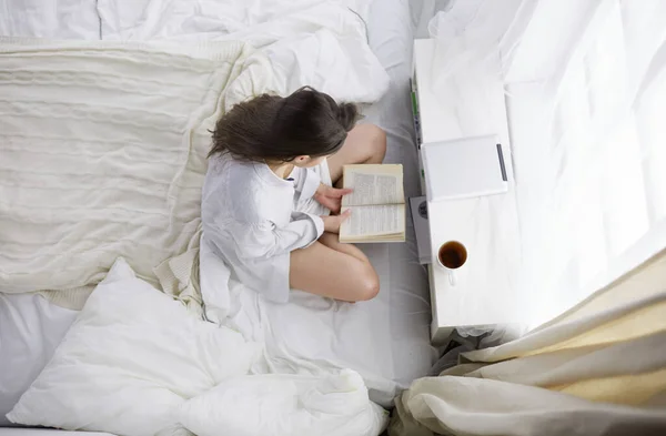 Mujer sentada en la cama leyendo un libro y desayunando — Foto de Stock