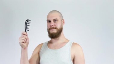 Çalı sakallı kel adam elinde tarak olan kameraya poz veriyor.