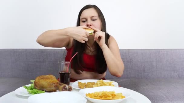 Spessa ragazza carina seduta sul divano e mangiare fast food, hamburger, patatine fritte . — Video Stock