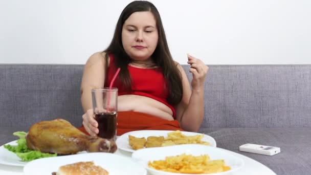 Pulchna kobieta siedząca przy stole i jedząca wysokokaloryczne niezdrowe jedzenie.Oglądanie telewizji. — Wideo stockowe