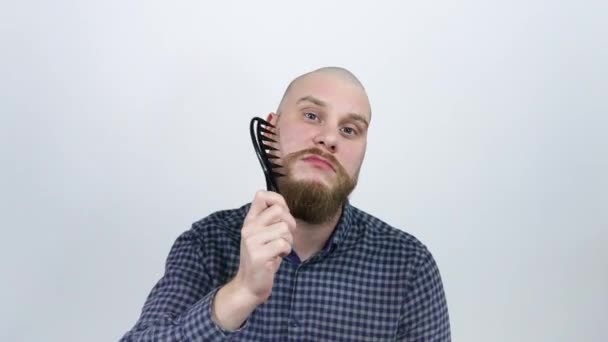 Мужчина с лысой головой расчесывает свою рыжую бороду и смотрит в камеру — стоковое видео