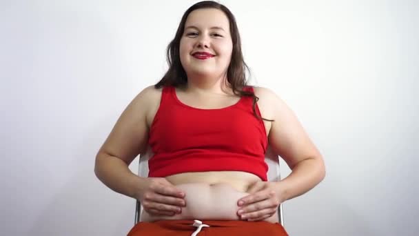 Dicke junge Frau sitzt auf einem Stuhl und berührt das Fett am Bauch. Lächelnd — Stockvideo