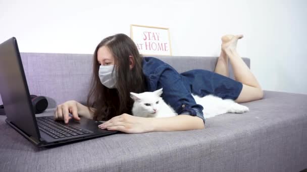 Женщина в медицинской маске работает дома на ноутбуке, а её кошка в медицинской маске — стоковое видео