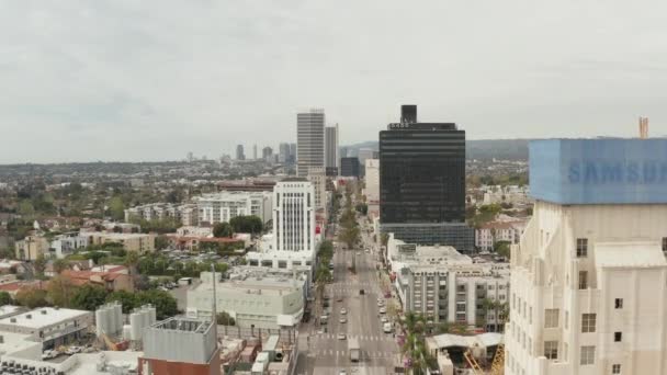 АЭРИАЛ: Полет над Уилширом Фабрегасом рядом с улицей и зданиями с автомобильным трафиком в Лос-Анджелесе, Калифорния в пасмурный день — стоковое видео