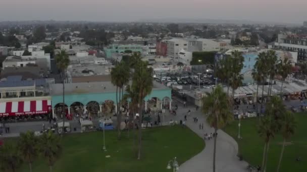 AERIAL: Venice Beach Boardwalkin yli vierailijoiden ja palmujen kanssa, Sunset, Los Angeles, Kalifornia — kuvapankkivideo