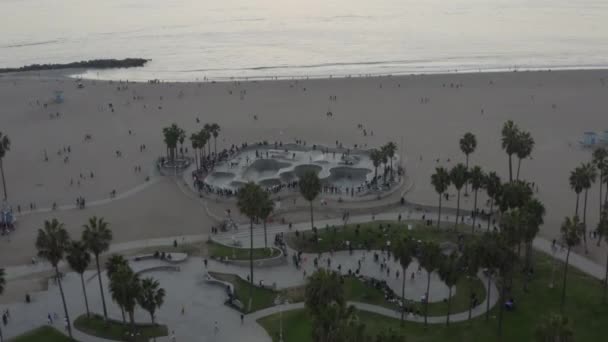 AERIAL: Volando lejos de Venice Beach Skatepark con visitantes y palmeras, puesta del sol en Los Ángeles — Vídeo de stock