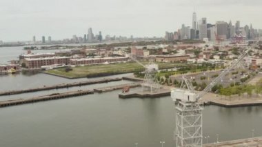 Rıhtımda, New York şehrinin arka planında ve nehrinde sanayi vinçleri daireler çiziyor. 