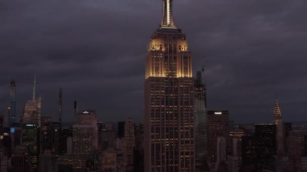 AERIAL: Voando até o Empire State edifício à noite em Manhattan cercado por arranha-céus em movimentada cidade — Vídeo de Stock