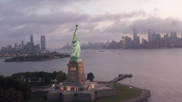 AERIELL: Cirklande Frihetsgudinnan upplyst tidigt på morgonen med dimmiga New York City Skyline i bakgrunden — Stockvideo