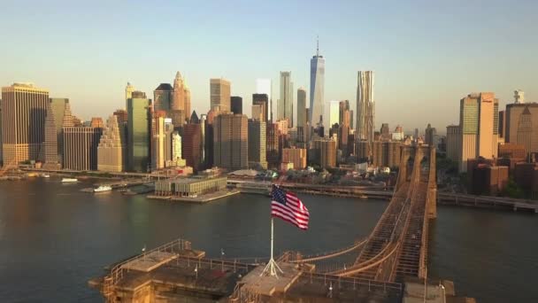 AERIAL: Полет над Бруклинским мостом с развевающимся американским флагом и видом на Ист-Ривер над Манхэттеном — стоковое видео