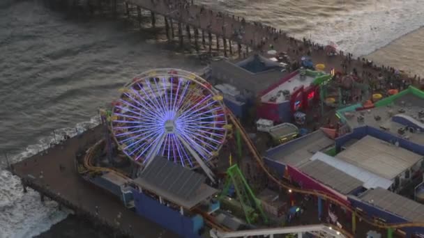 AERIAL: Circling Santa Monica Pier, Los Angeles shora při krásném západu slunce s turisty, chodci baví v zábavním parku Ferrys Wheel s výhledem na oceán vlny shazovat — Stock video