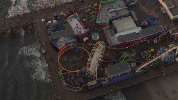 AERIAL: Círculo de Santa Monica Pier, Los Angeles a partir de cima no belo pôr do sol com turistas, pedestres se divertindo no parque temático Ferrys Roda com ondas vista mar bater — Vídeo de Stock