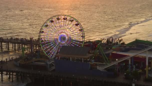 AERIAL: Blisko widok Santa Monica Pier Ferrys Wheel, Los Angeles na piękny zachód słońca z turystami, pieszych spacerujących bawiąc się w parku rozrywki rollercoaster z widokiem na ocean fale awarii — Wideo stockowe