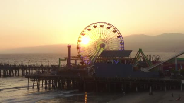 AERIAL: Vista de cerca de Santa Monica Pier Ferrys Wheel, Los Ángeles en el hermoso atardecer con turistas, peatones caminando divirtiéndose en la montaña rusa del parque temático con olas de vista al mar estrellándose — Vídeos de Stock