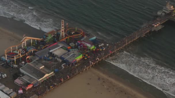 AERIAL: Cirkling Santa Monica Pier, Los Angeles från ovan vid vackra solnedgången med turister, Fotgängare som har roligt på nöjespark Ferrys Wheel med havsutsikt vågor kraschar — Stockvideo