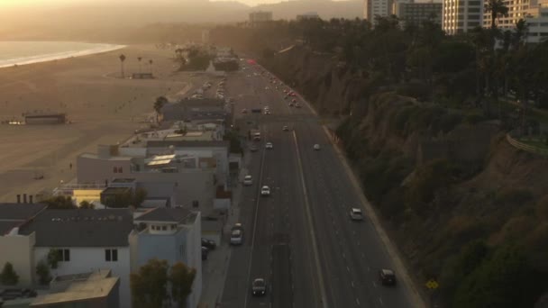 AERIAL: Uitzicht op Pacific Coast Highway PCH naast Santa Monica Pier, Los Angeles met licht verkeer en uitzicht op de oceaan door bij zonsondergang golven, zomer — Stockvideo