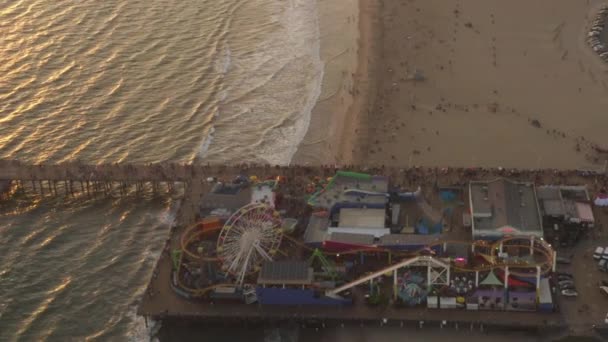 エリアル:ロサンゼルスのサンタモニカ桟橋から観光客と一緒に美しい日没で飛んで、海の景色の波がクラッシュするテーマパークジェットコースターで楽しんで歩く歩行者 — ストック動画