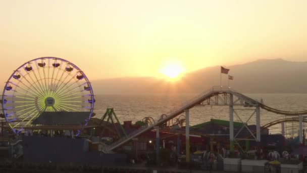 AERIAL: Vista de cerca de Santa Monica Pier Ferrys Wheel, Los Ángeles en el hermoso atardecer con turistas, peatones caminando divirtiéndose en la montaña rusa del parque temático con olas de vista al mar estrellándose — Vídeo de stock