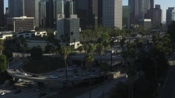 AERIAL: W kierunku centrum Los Angeles, California skrzyżowania ruchu z palmami i Skyline w tle w pięknym błękitnym niebie i słoneczny dzień — Wideo stockowe