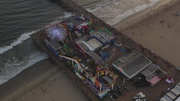 AERIAL: körözés Santa Monica móló, Los Angeles felülről gyönyörű naplemente a turisták, gyalogosok szórakozás vidámpark Ferrys Wheel óceánra néző hullámok összeomlik — Stock videók