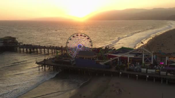 AERIAL: Blisko widok Santa Monica Pier Ferrys Wheel, Los Angeles na piękny zachód słońca z turystami, pieszych spacerujących bawiąc się w parku rozrywki rollercoaster z widokiem na ocean fale awarii — Wideo stockowe