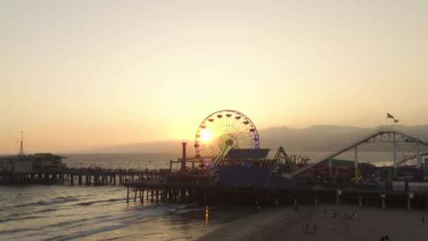 AERIAL: Close-up uitzicht op Santa Monica Pier Ferrys Wheel, Los Angeles bij prachtige zonsondergang met toeristen, voetgangers wandelen plezier op pretpark achtbaan met uitzicht op de oceaan golven crashen — Stockvideo