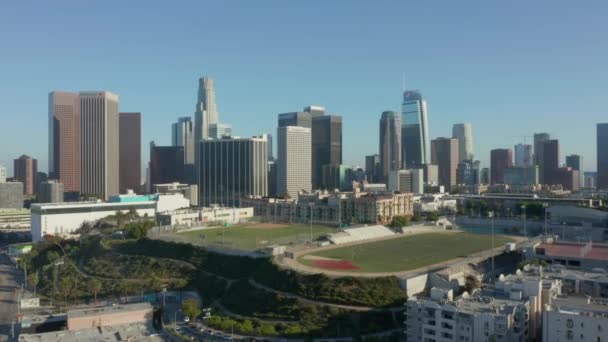 AERIAL: Volando hacia el centro de Los Ángeles, California Skyline con béisbol de fútbol deportivo y campo de tenis en el hermoso cielo azul y día soleado — Vídeo de stock