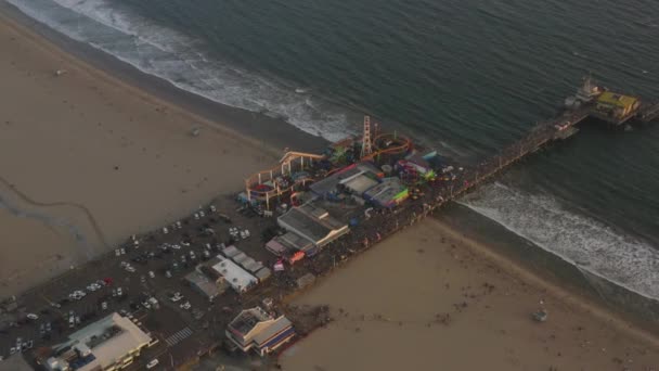 AERIAL: Circling Santa Monica Pier, Los Angeles van boven bij prachtige zonsondergang met toeristen, Voetgangers hebben plezier op pretpark Ferrys Wiel met uitzicht op de oceaan golven crashen — Stockvideo