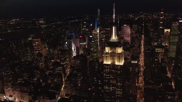 AERIAL: dech beroucí pohled na ikonickou Empire State Building nahoře osvětlené paralelní cesty a křižovatky obytných bytů a kancelářských budov v Midtown Manhattan, New York City v noci — Stock video