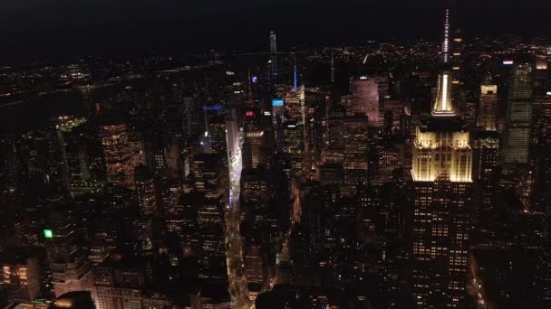 AERIAL: dech beroucí pohled na ikonickou Empire State Building nahoře osvětlené paralelní cesty a křižovatky obytných bytů a kancelářských budov v Midtown Manhattan, New York City v noci — Stock video